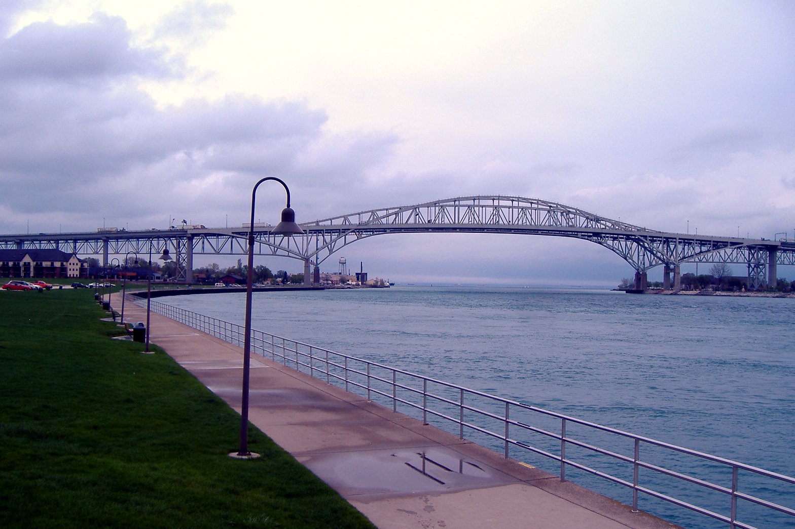 A paved riverside walkway with a railing and lamp posts runs along the St. Clair River toward the Blue Water Bridge under a gray sky.