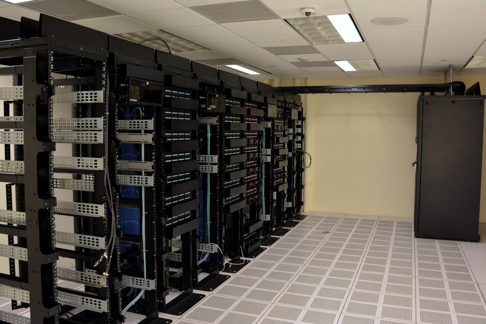 Rows of black server racks with blue network cables stand on a raised, perforated floor inside a data center room, in a file photo.