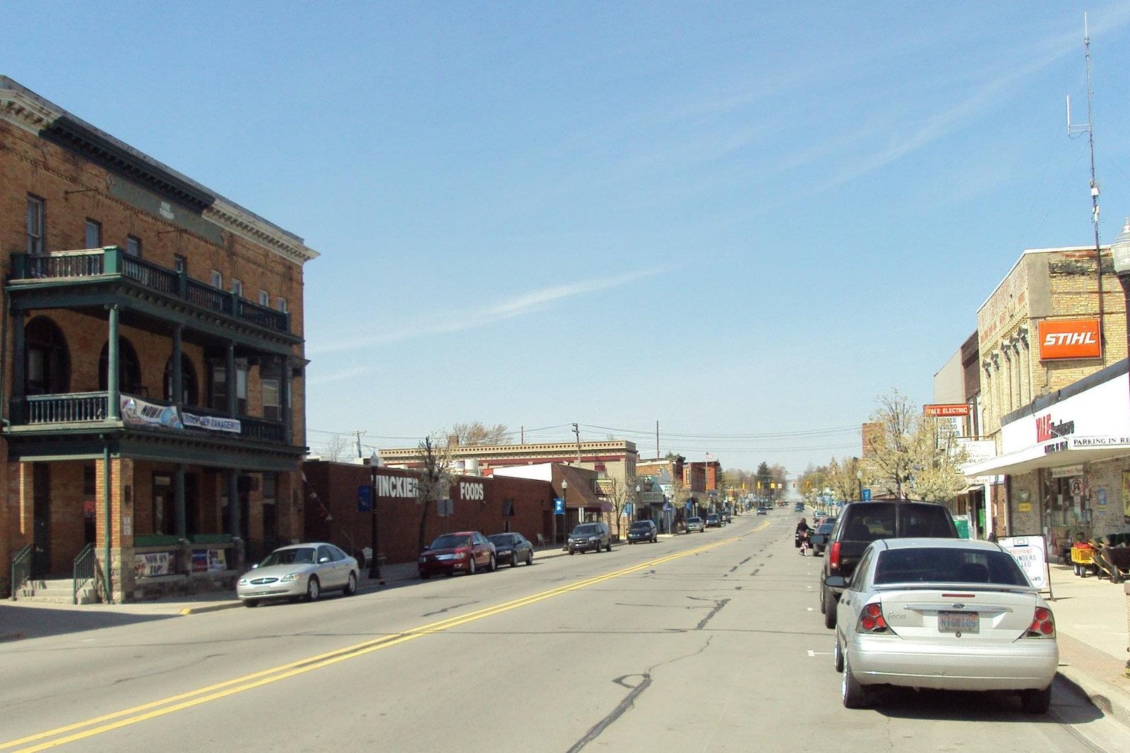 Downtown Yale, Michigan, looking down Main Street on a sunny day, with a two-story brick building with green balconies on the left, storefronts on both sides and cars parked along the curb.