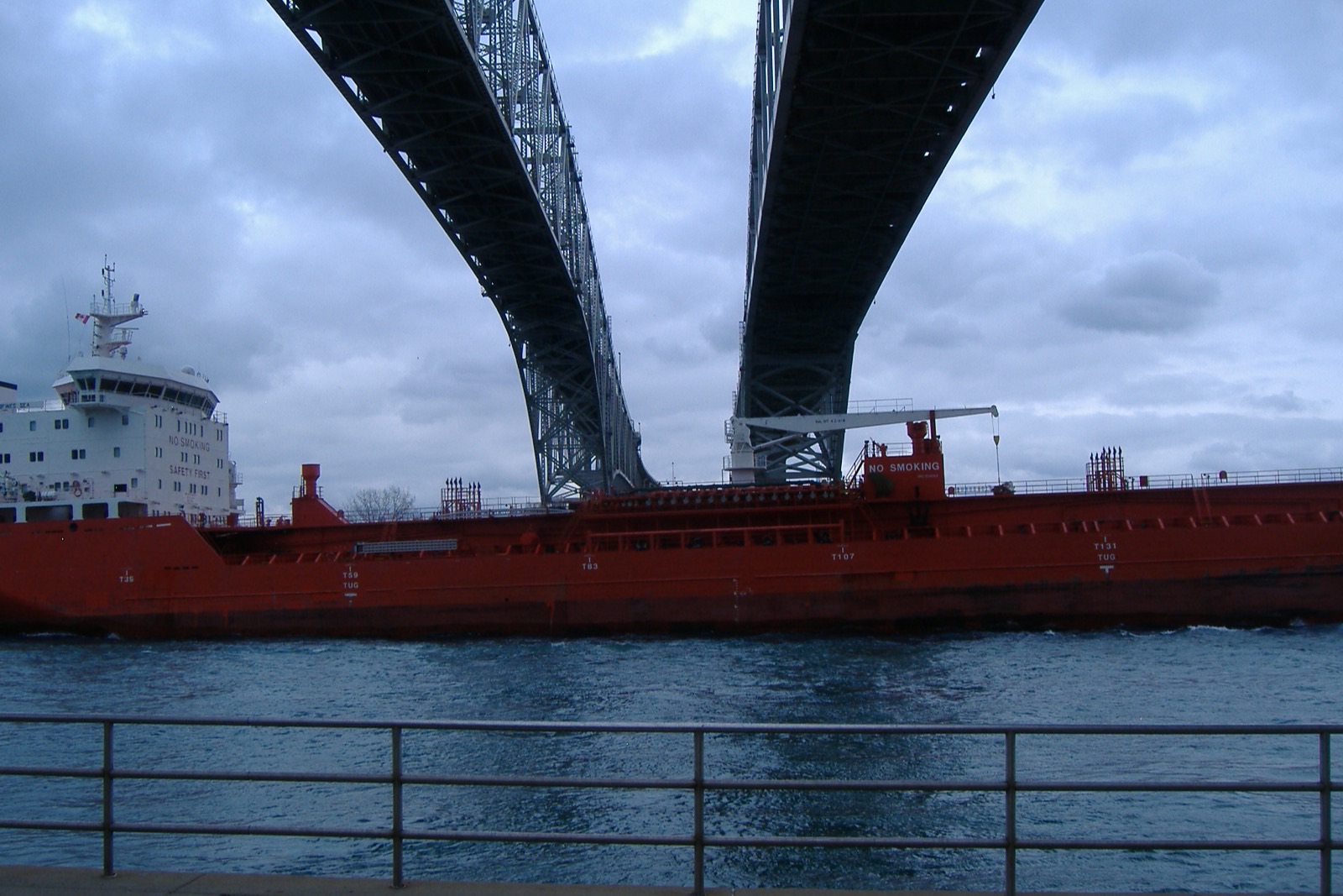 A red tanker passes beneath the twin spans of the Blue Water Bridge at Port Huron, seen from a riverfront railing.