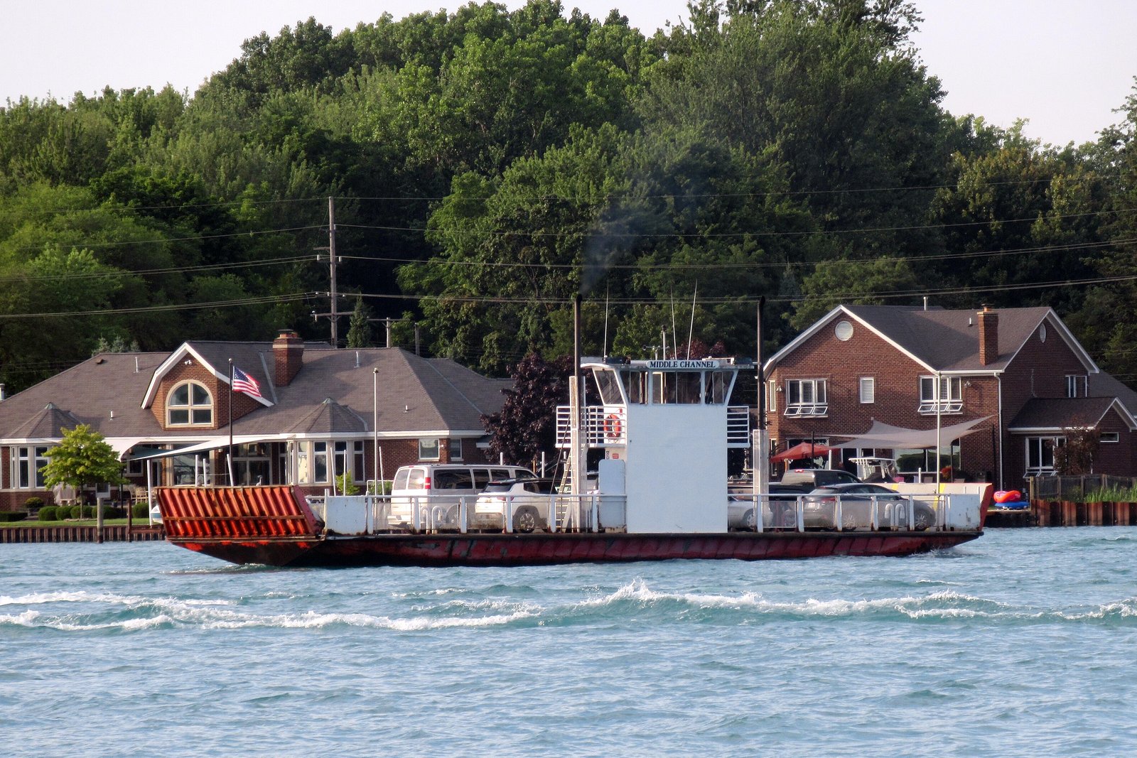 A small car ferry with a red-and-white striped ramp crosses a river past brick houses and tall trees.