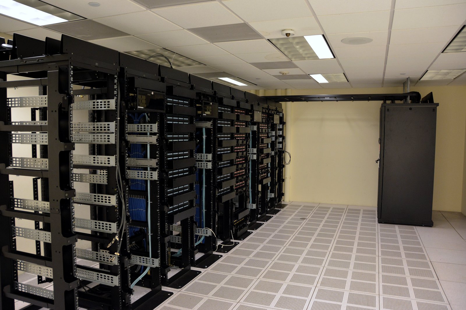 Rows of black open-frame server racks with patch cables stand on a perforated raised floor inside a data center room.