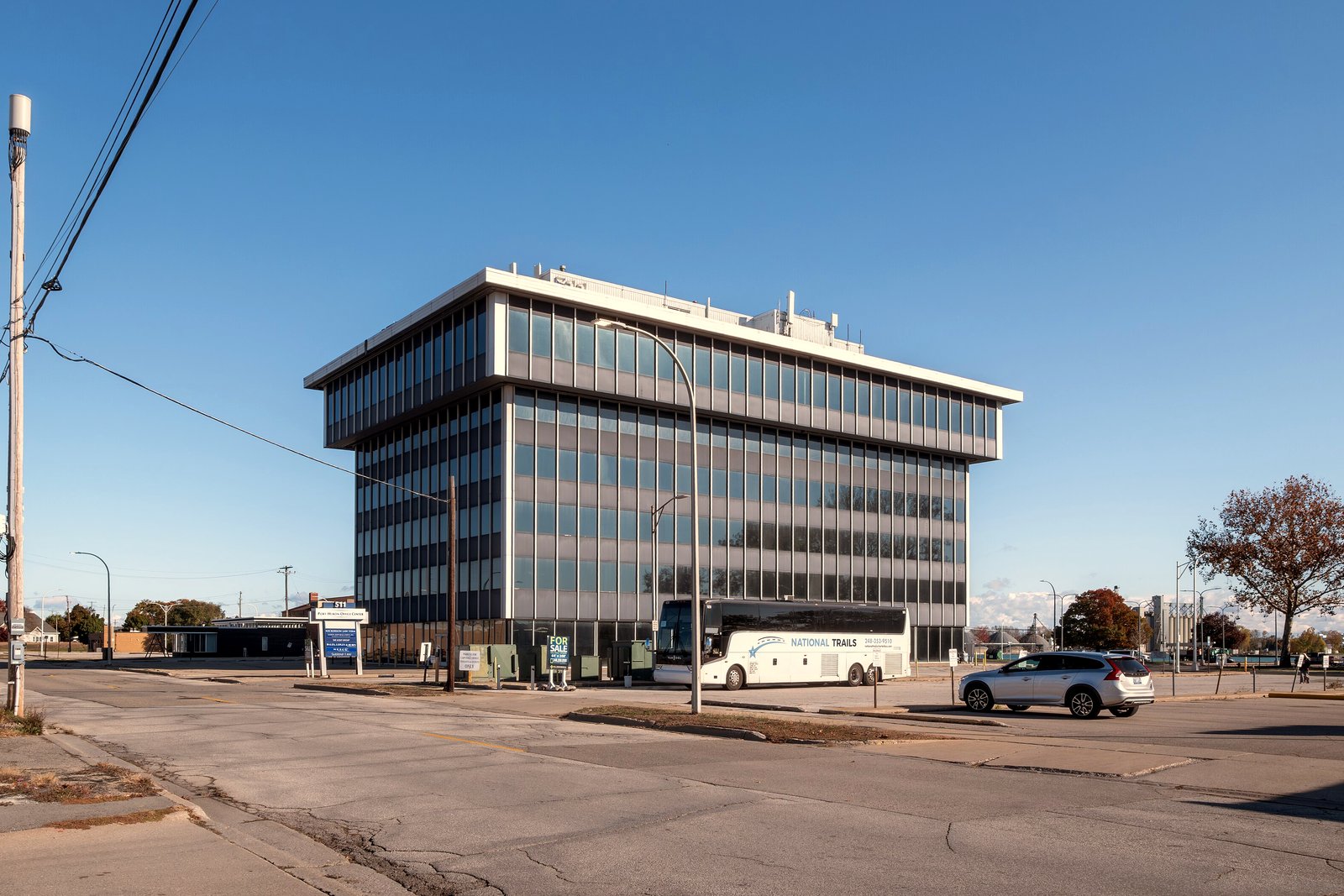A modern multistory office building with rows of glass windows beside a wide, nearly empty street and parking lot in downtown Port Huron under a clear blue sky.