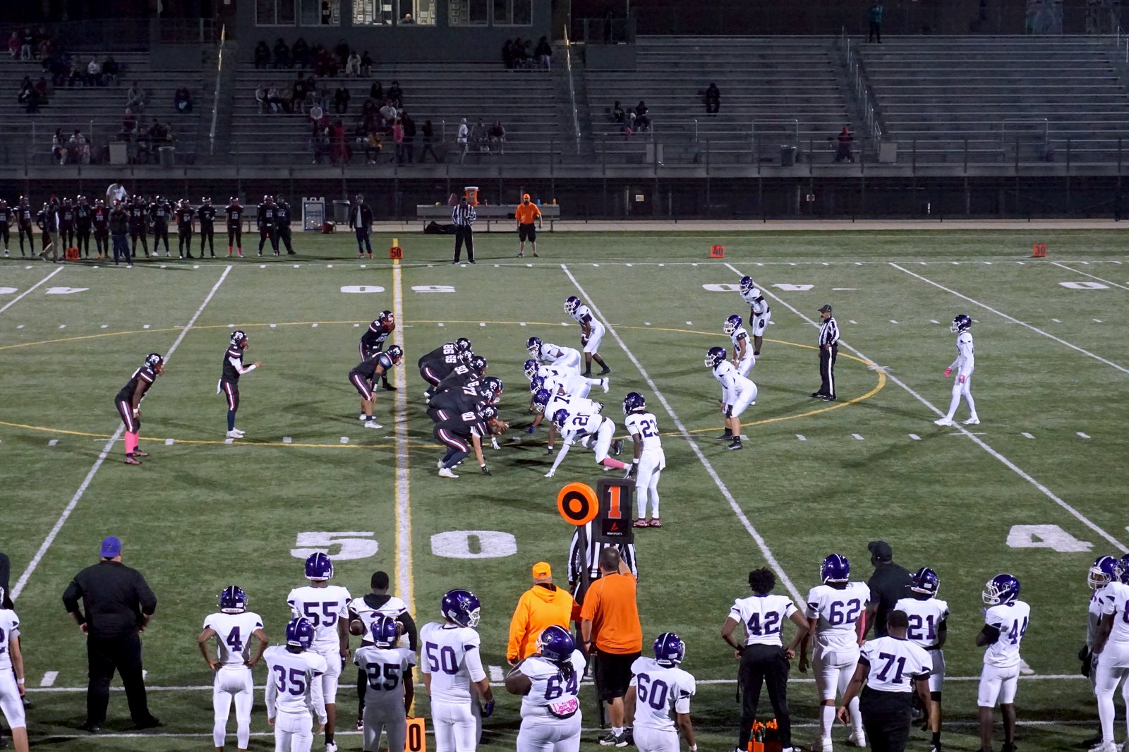 High school football players in dark and white uniforms face off at the line of scrimmage on a lit night field, with white-jersey sideline players in the foreground near midfield.