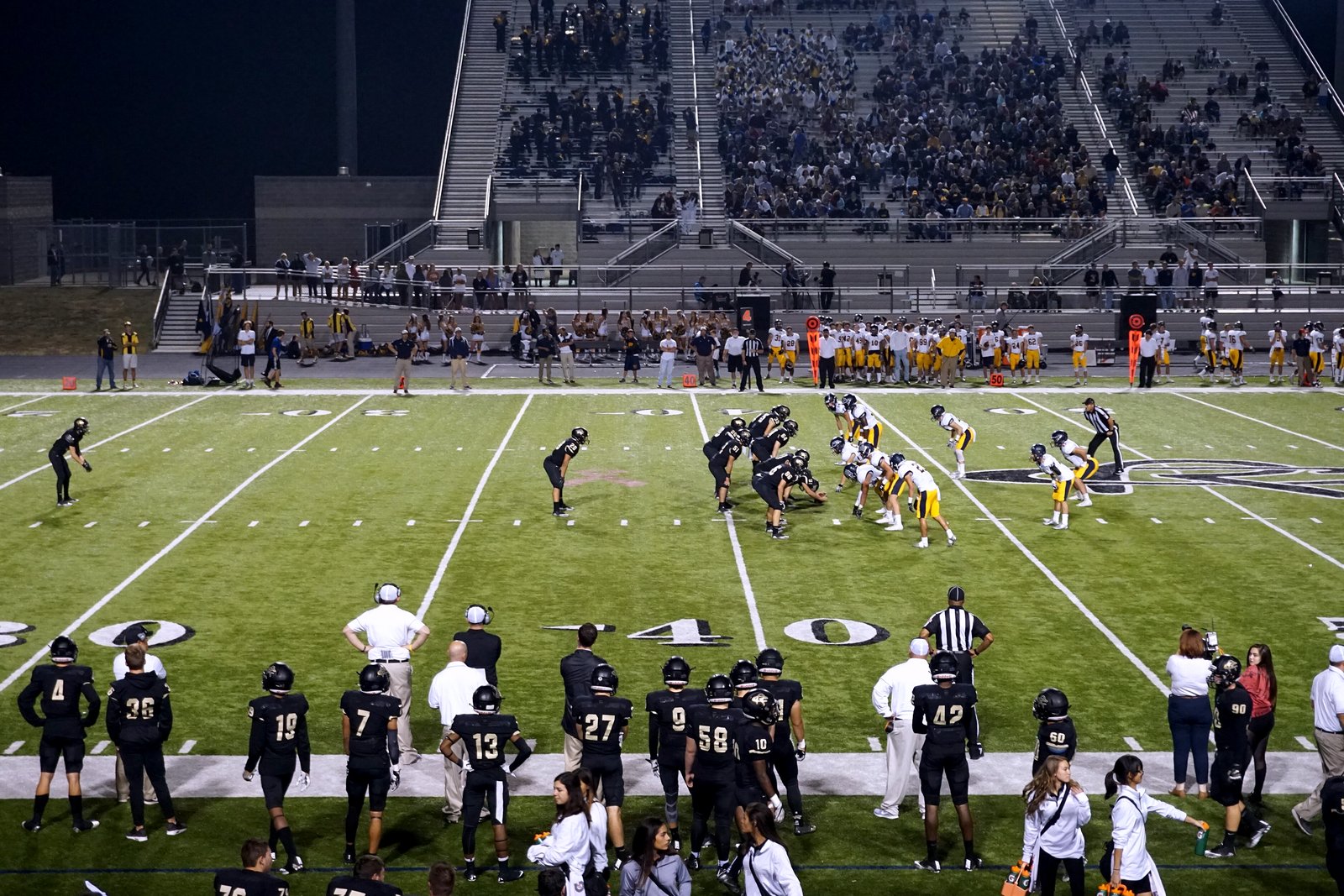 A high school team in dark jerseys watches from the sideline as players on a lit turf field line up for a punt, with a grandstand behind them.