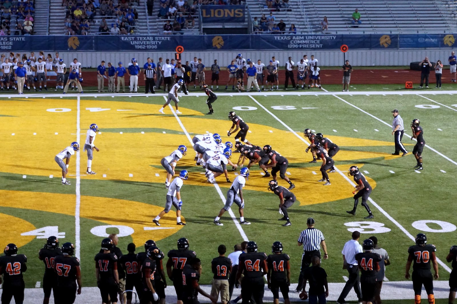 High school football players in dark and white uniforms battle at the line of scrimmage during a daytime game.