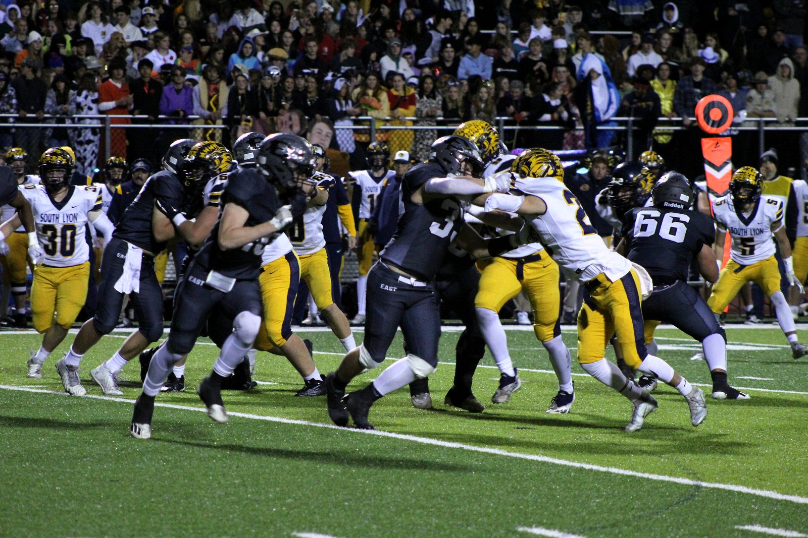 High school football players in black and white and in yellow and white jerseys collide at the line of scrimmage on a lit field, with a crowd in the stands behind them.