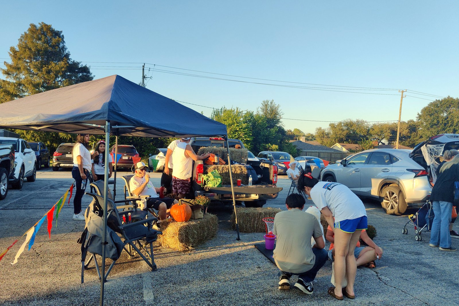Families gather under a blue canopy and around decorated cars with open trunks in a parking lot, with hay bales and pumpkins, in late-afternoon sun.
