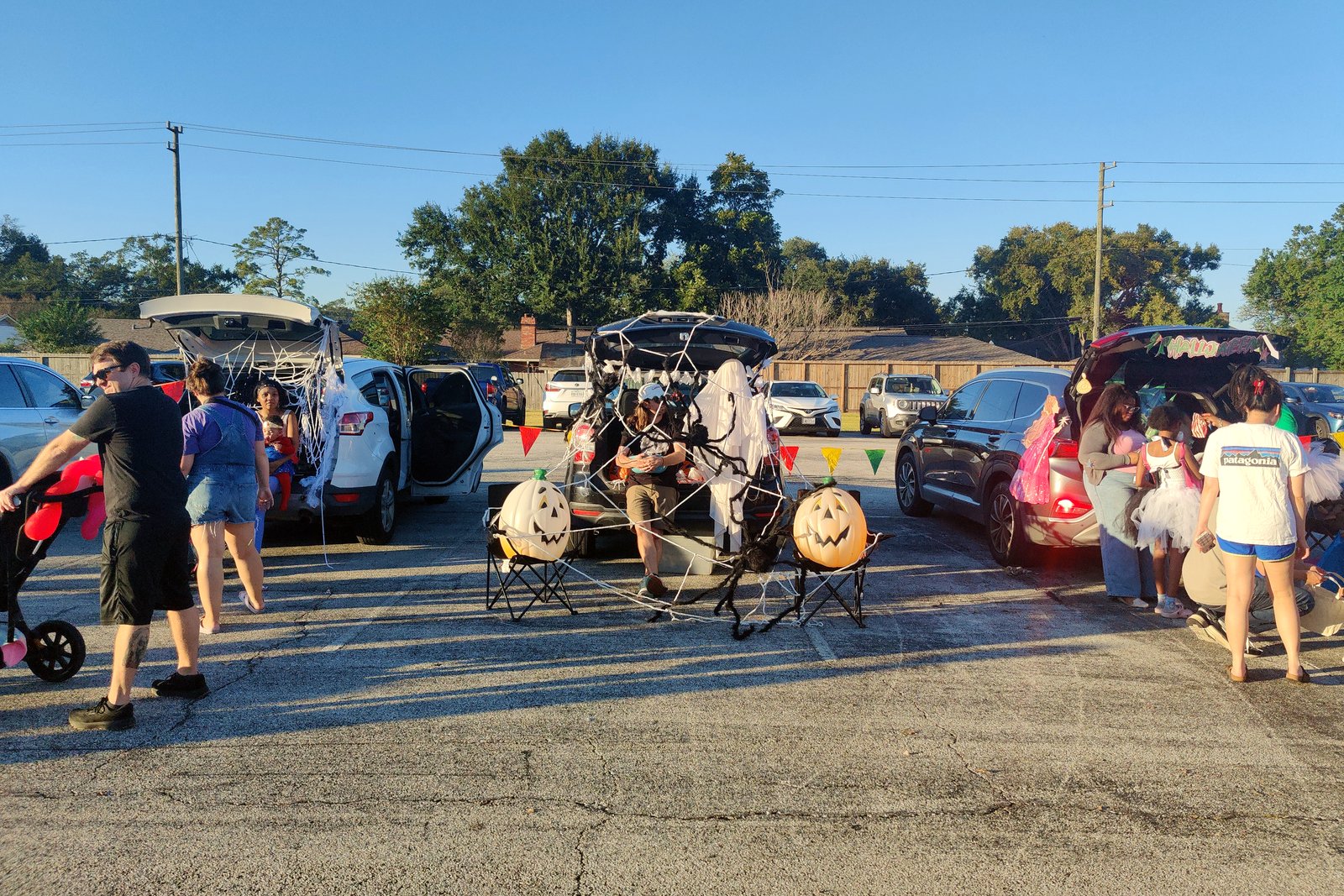 Families and children in costumes walk past cars with open trunks decorated for Halloween in a parking lot on a sunny afternoon.