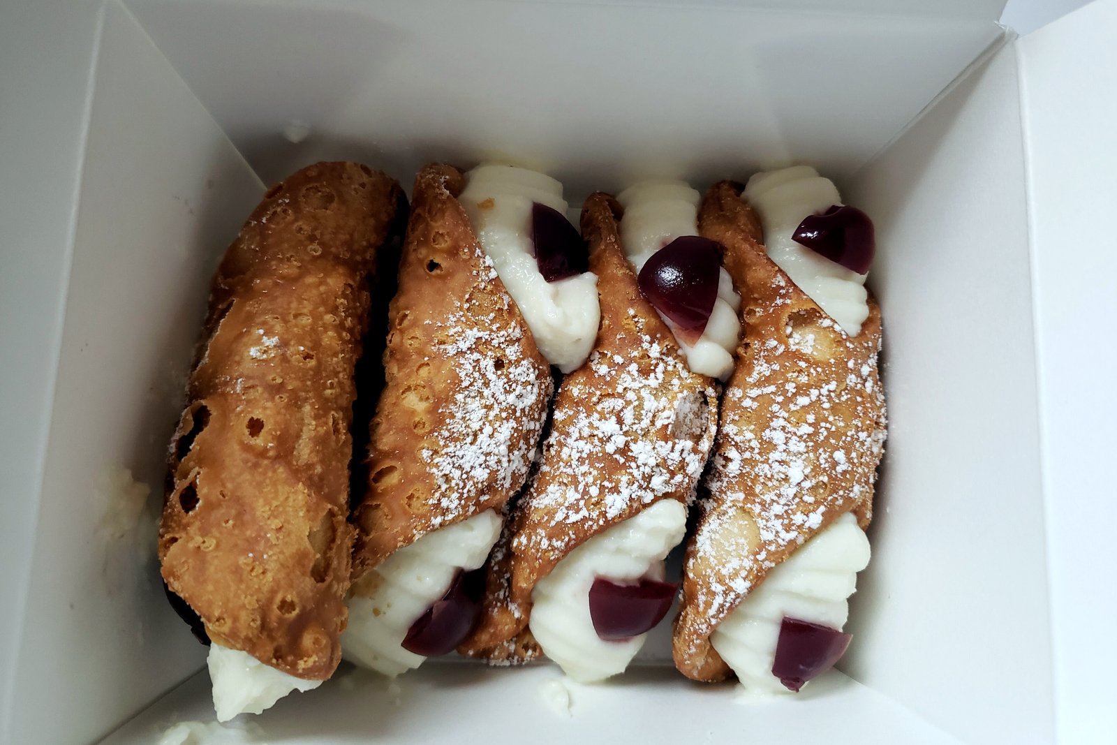 Four cannoli dusted with powdered sugar, with cherries at the ends, sitting in an open white bakery box.