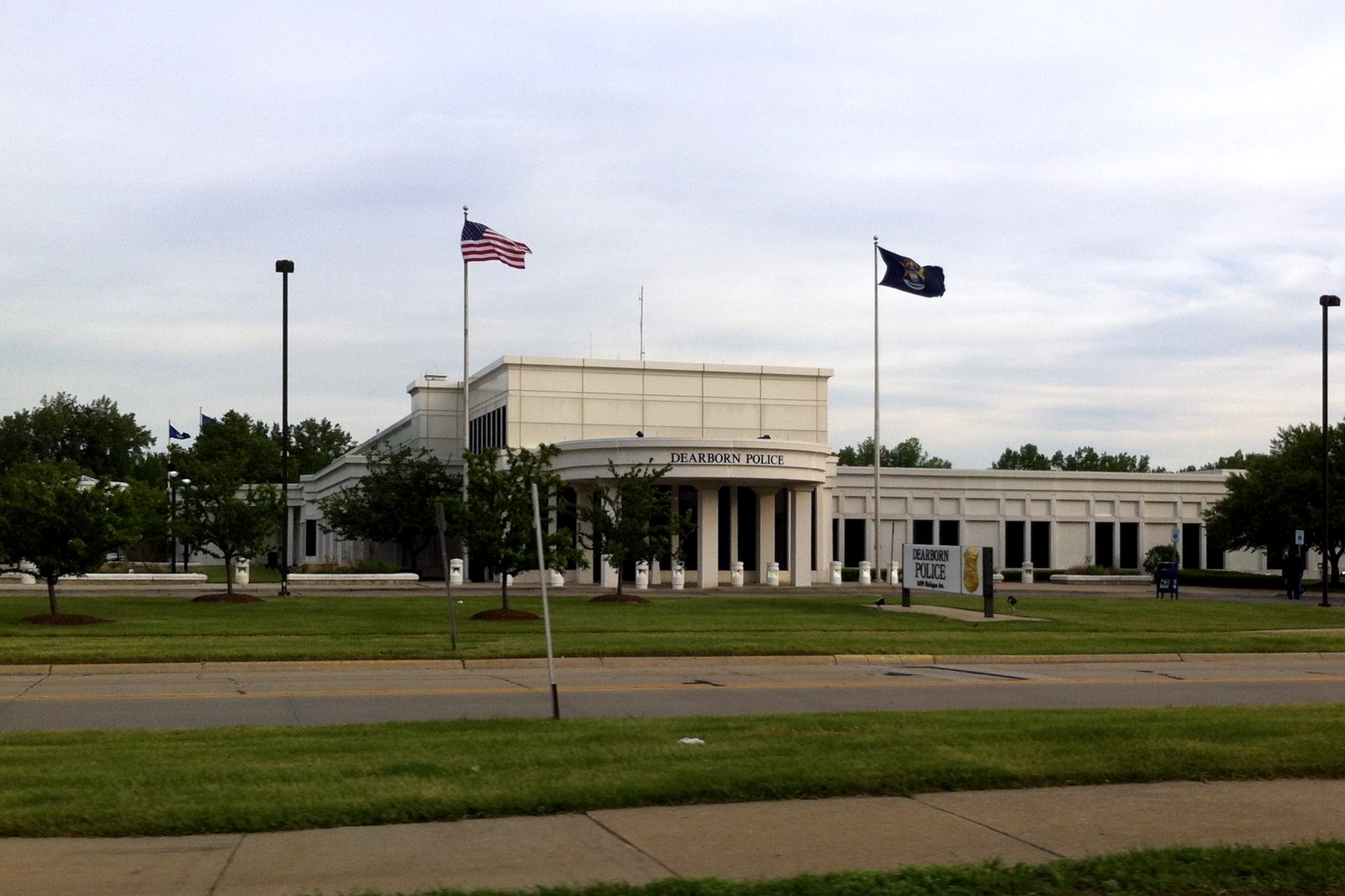 A large white classical-style building with columns and two flagpoles flying an American flag and a dark blue flag behind a wide green lawn.