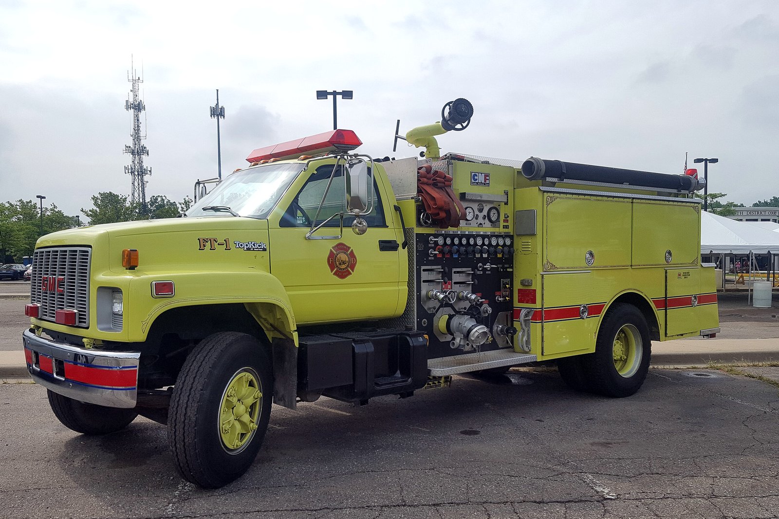 A yellow fire engine with chrome bumpers and red lights parked on a paved lot under a cloudy sky.