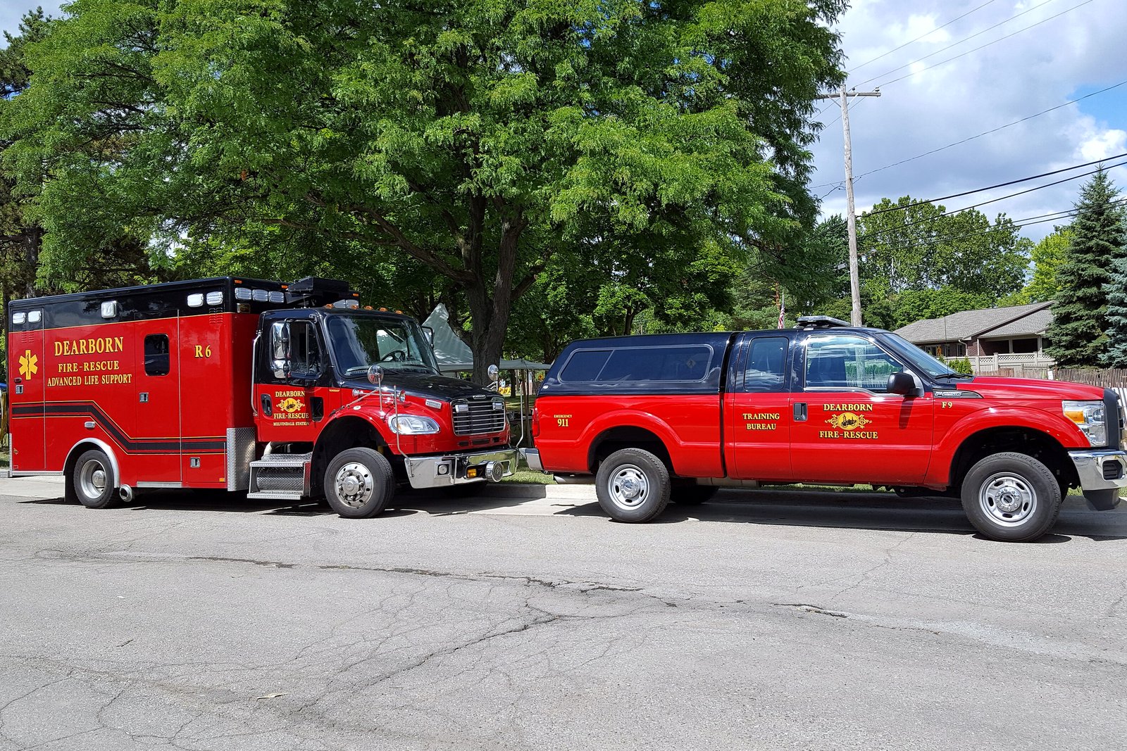 A red fire department ambulance and a red pickup truck parked side by side on pavement in front of green trees.