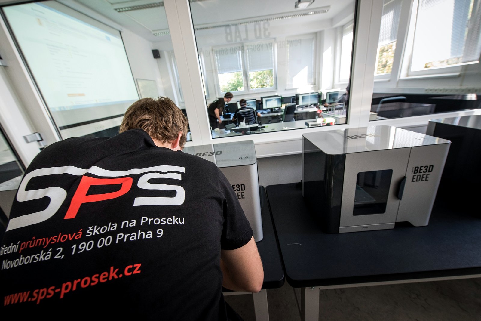 A student in a black school T-shirt works at a desk in a glass-walled classroom with several small white 3D printers.