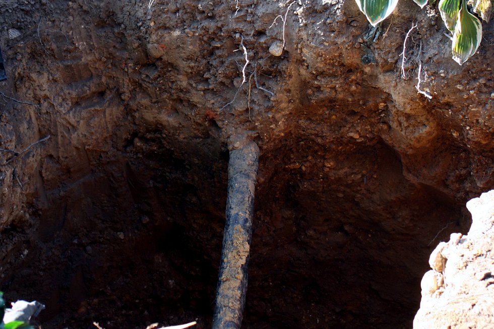 A deep hole dug in a yard next to plants, with a gray lead water pipe running down through the dirt.