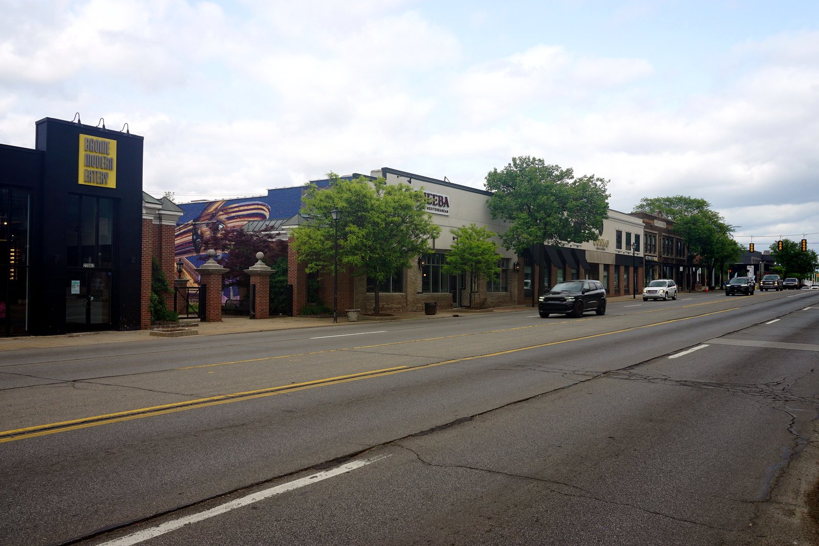 A wide, multi-lane street lined with low brick storefronts and young trees, with a few cars in the distance under a partly cloudy sky.