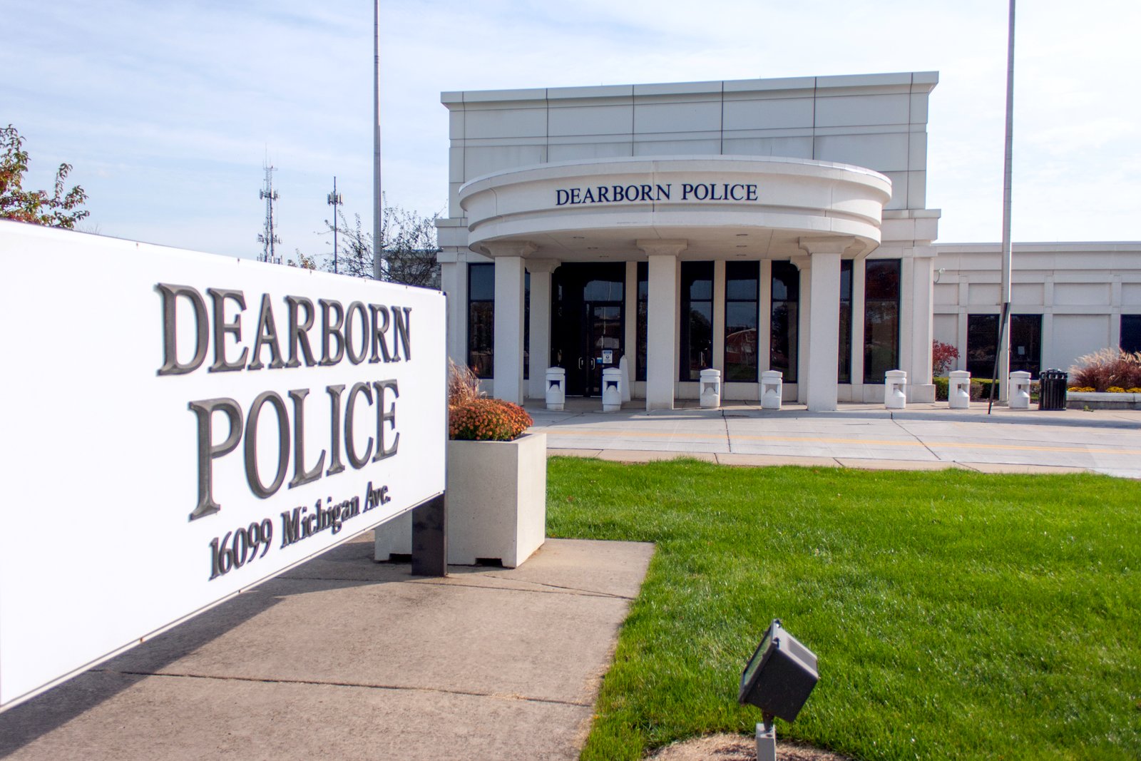 A white stone police building with a curved facade behind a large white sign reading Dearborn Police, with a lawn and planters in front.