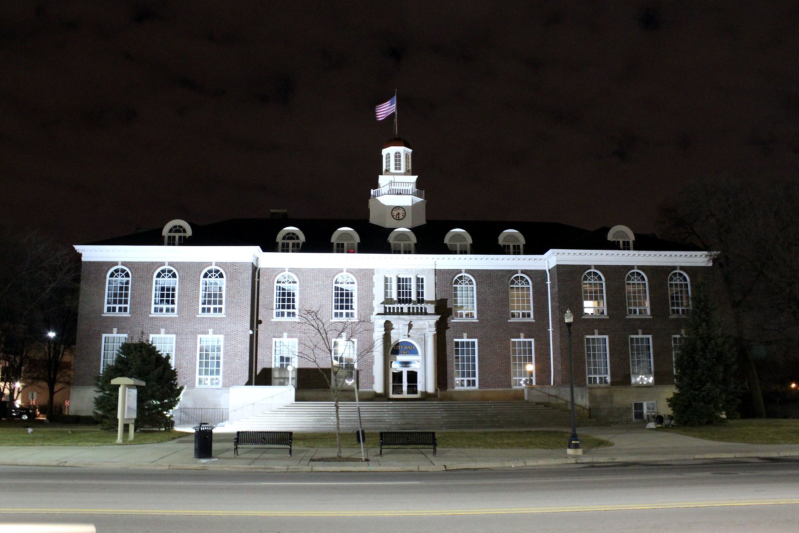 A two-story brick civic building with white trim, a white cupola and a flag on top, lit up at night.