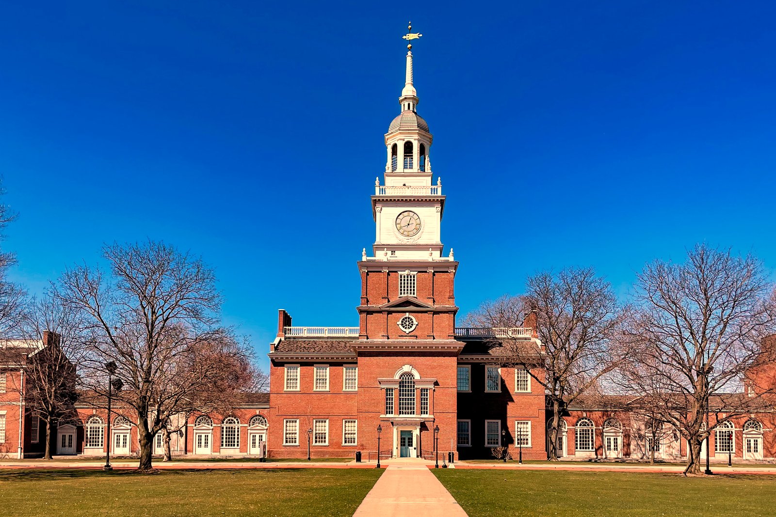 A red brick building with a white clock tower and steeple behind a wide brick walkway and bare trees under a blue sky.