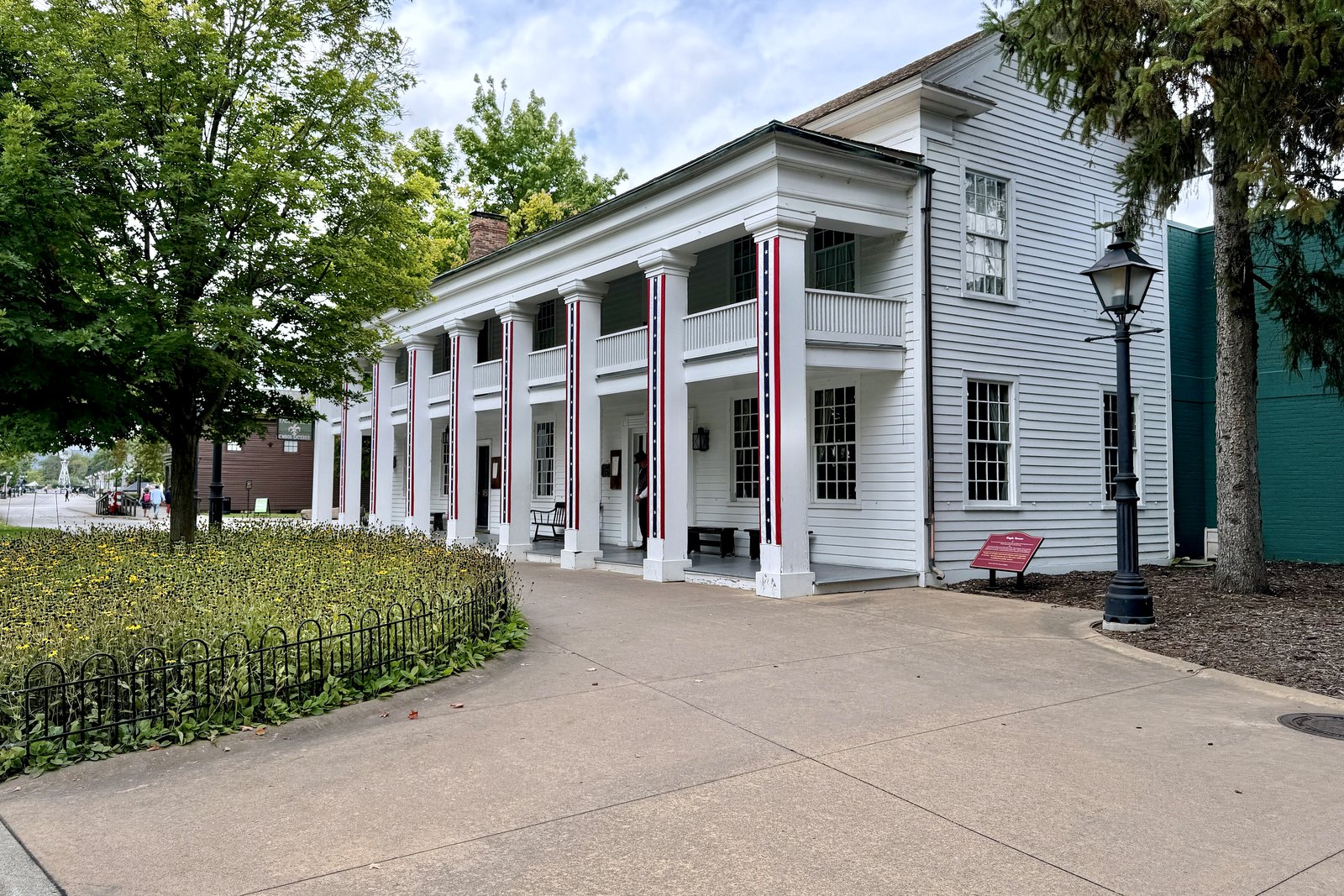 A white two-story Greek Revival building with a long columned porch, framed by trees and a lawn under a blue sky.