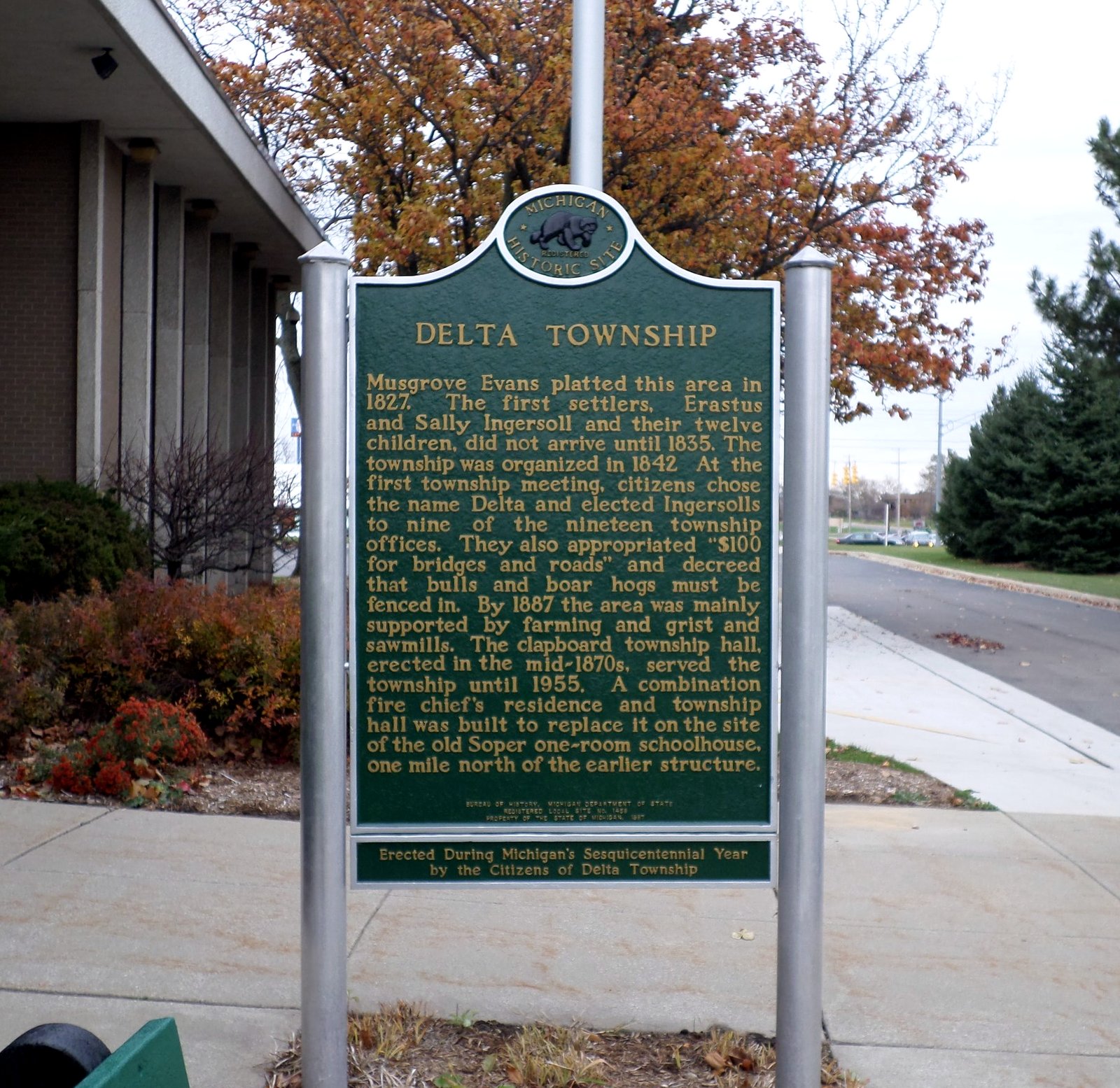 A green Michigan historical marker titled Delta Township stands on two posts beside a sidewalk, with a building and autumn trees behind it.