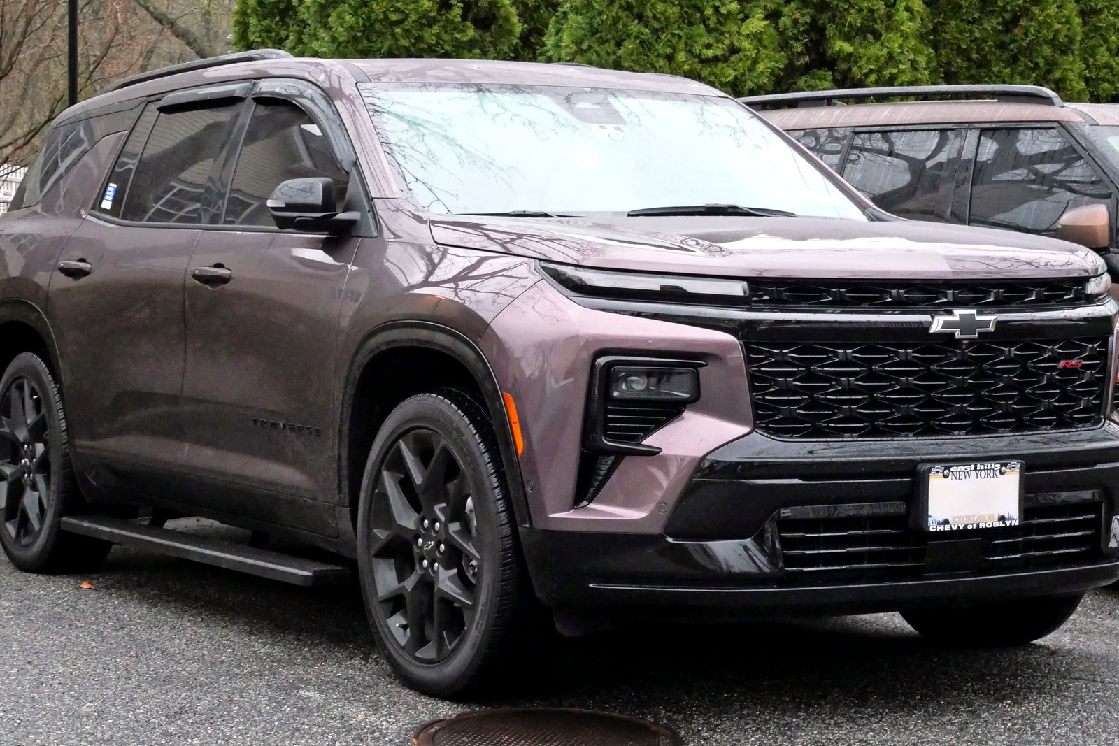 A dark mauve Chevrolet Traverse RS SUV with a black grille and black wheels, parked in a lot in front of evergreen shrubs.