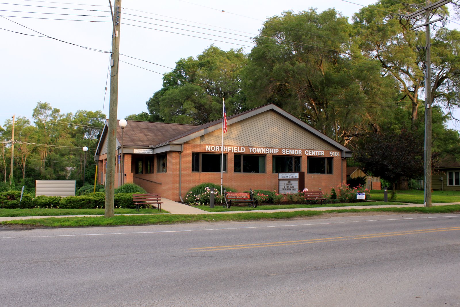 A one-story brick senior center with a peaked roof, a flagpole, benches and flower beds along a two-lane road.