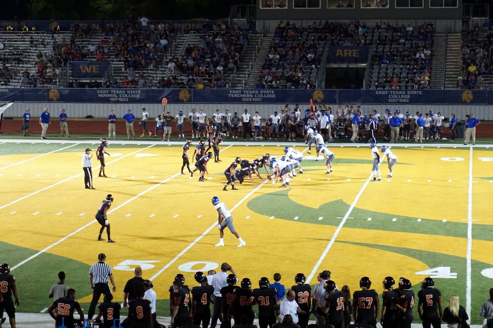 High school football players line up at the line of scrimmage on a gold-and-green field at night as teammates watch from the sideline.