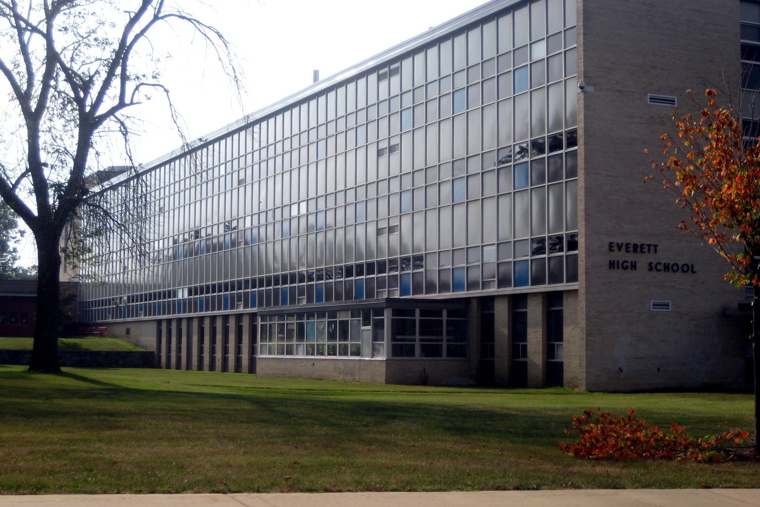 A long, modern school building with rows of tall windows and a lettered sign reading Everett High School, beside a lawn and a leafless tree.