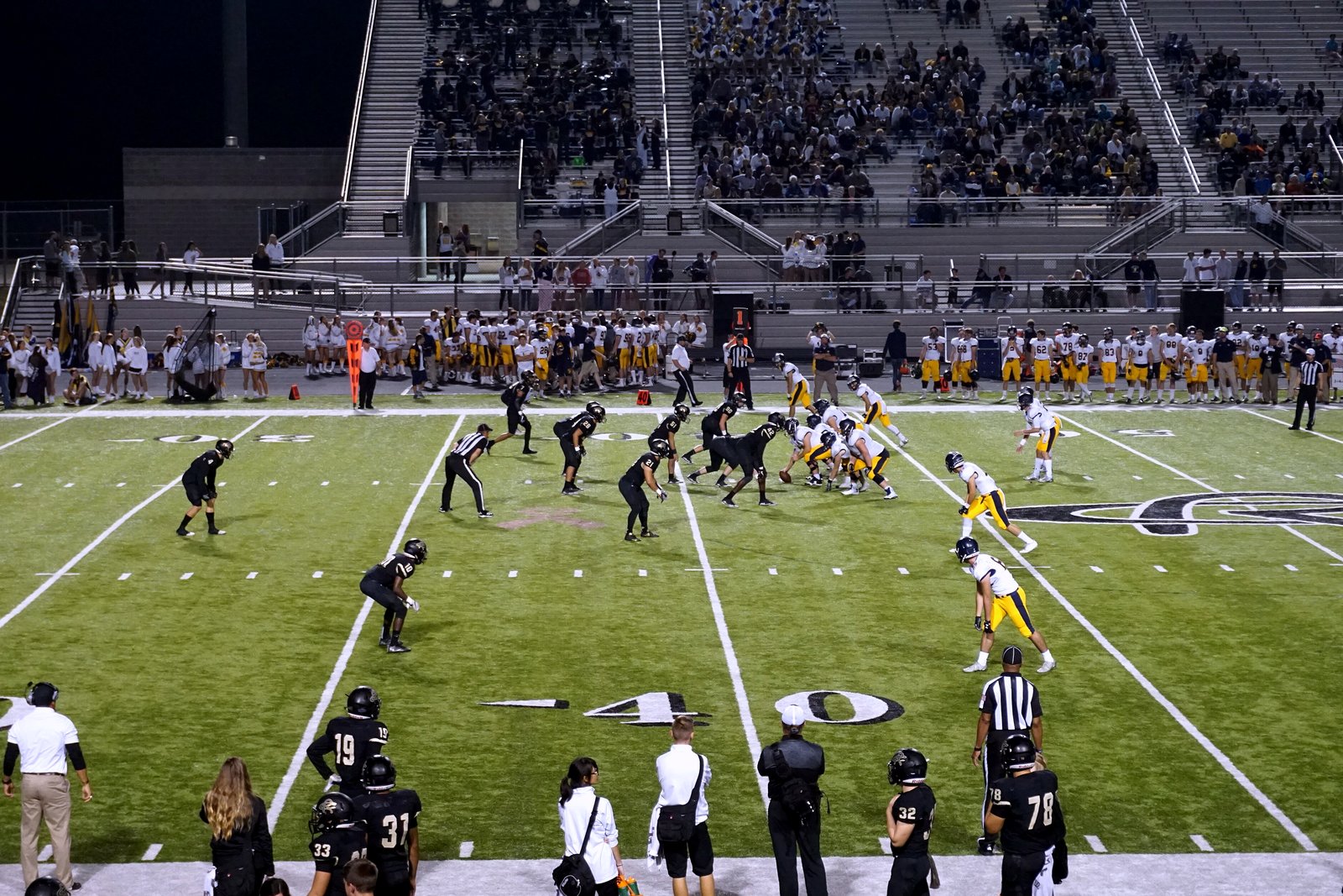 A high school football team in white and gold lines up against a team in black at the line of scrimmage on a lighted field at night, with fans in the stands.