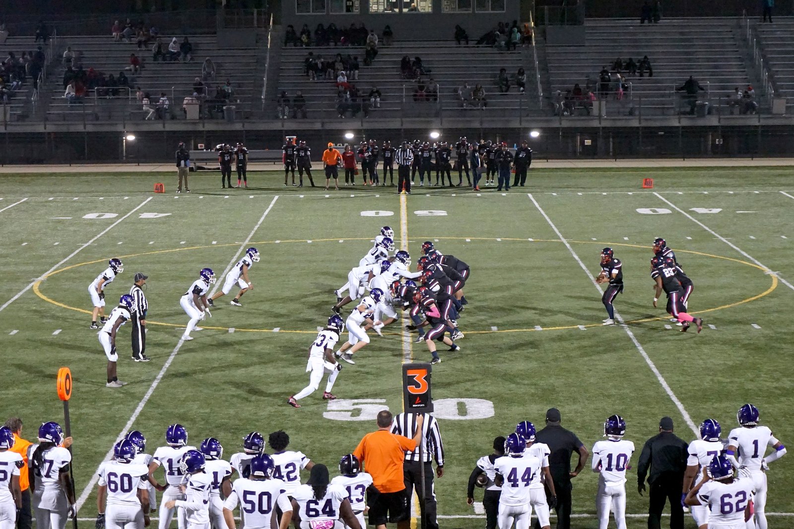 Two high school football teams face off at the line of scrimmage on a turf field, with players in white and purple watching from the sideline in front.