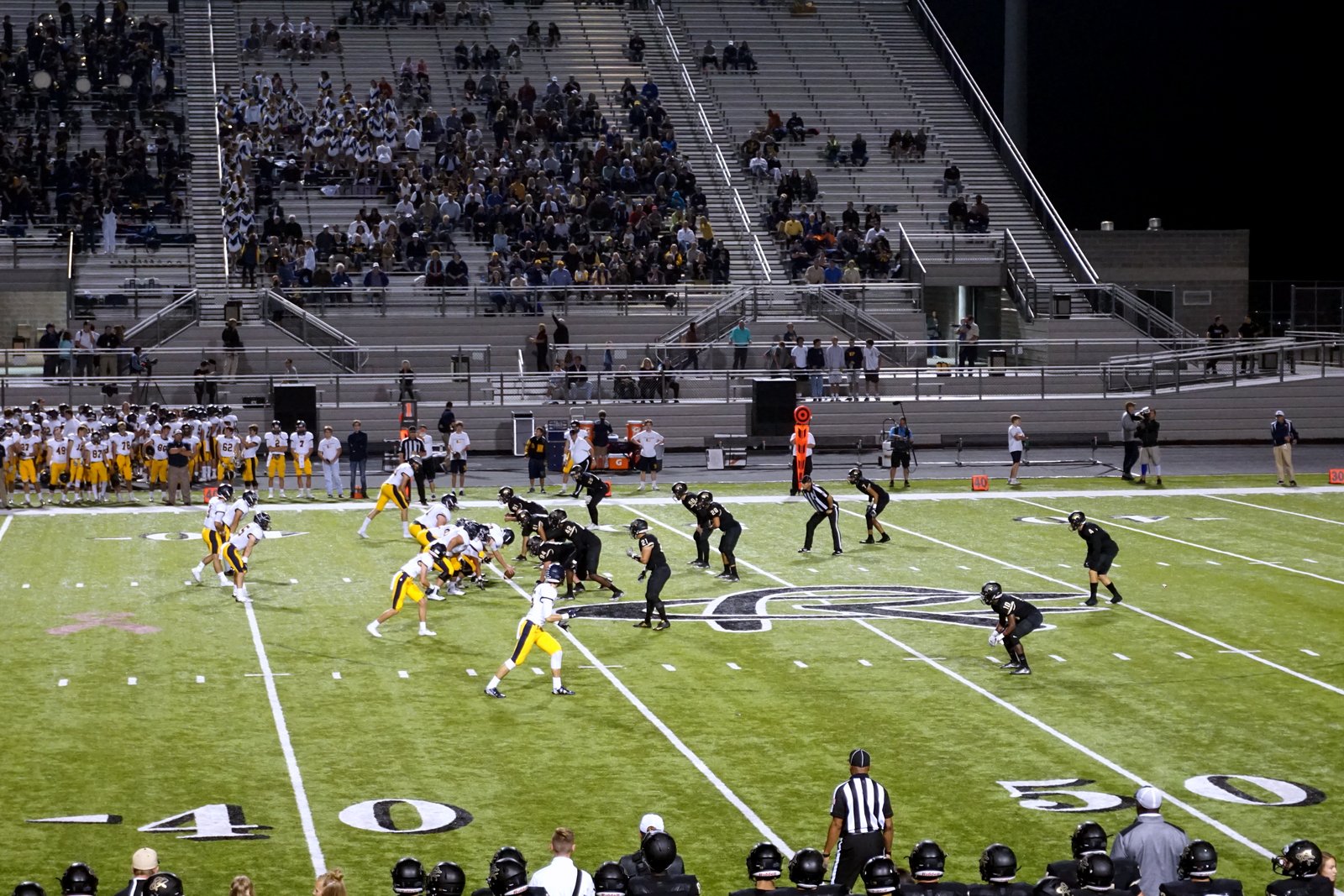 High school football players in white and gold line up on offense against a team in black at night, with a crowd in the stadium bleachers behind them.