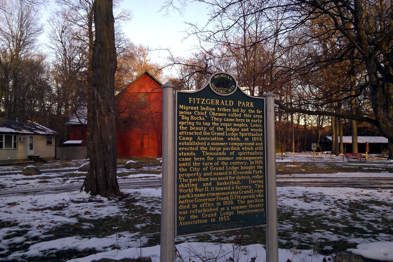 A green Michigan historical marker for Fitzgerald Park stands among bare trees on a lawn patched with snow, with a red building behind it.