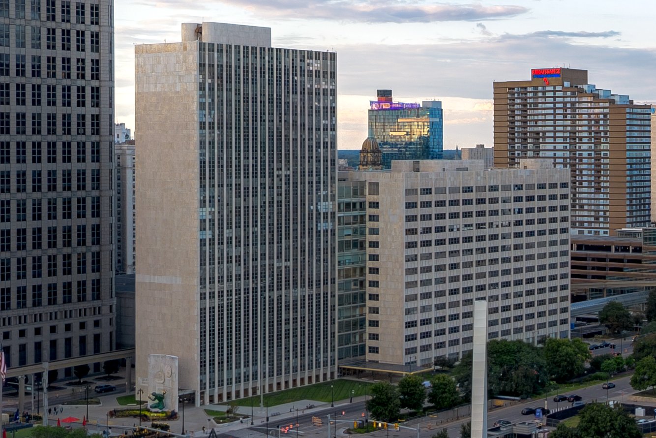 A tall pale municipal tower and a lower wing beside a plaza with a green sculpture, with downtown Detroit buildings behind under an evening sky.