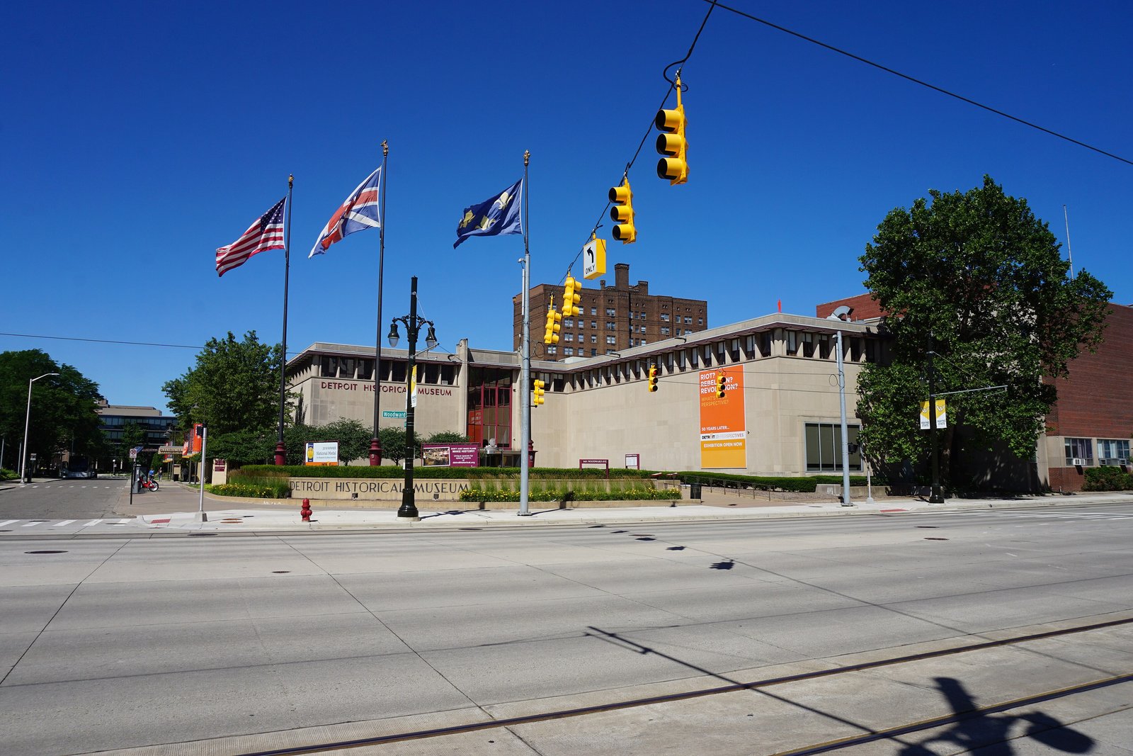 A low modern stone museum building with tall windows along a tree-lined street in Detroit’s Midtown under a blue sky.