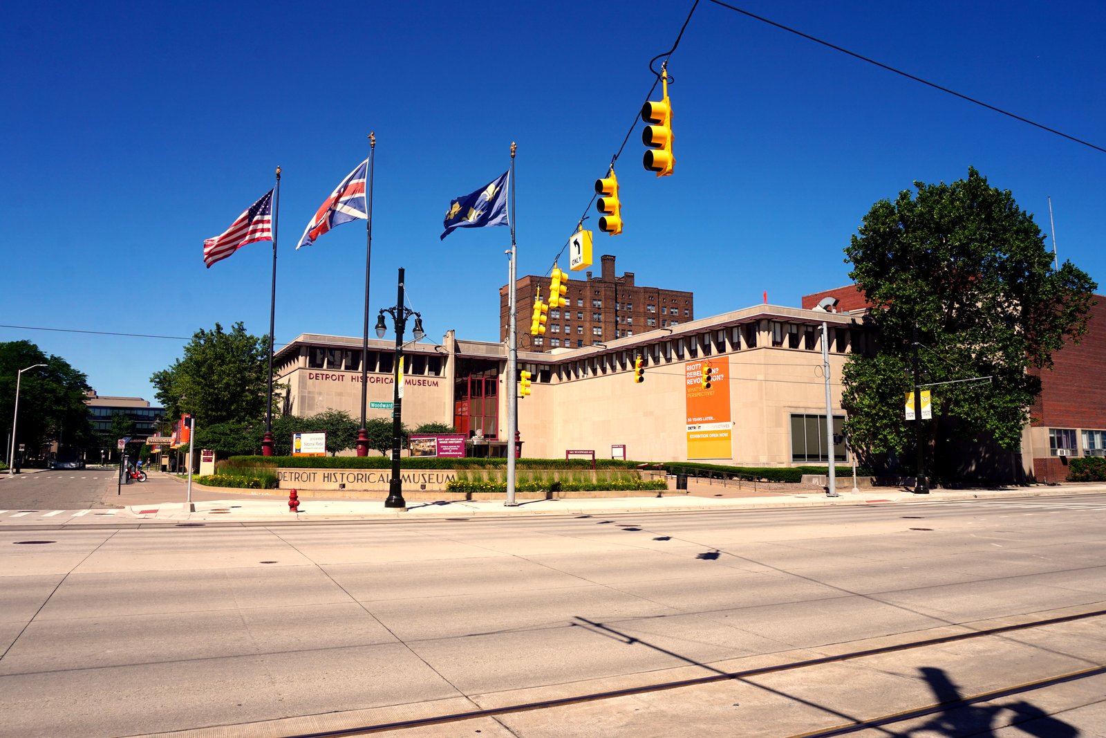 The low, tan stone Detroit Historical Museum sits behind a wide, empty Woodward Avenue intersection with flags flying out front under a bright blue sky.