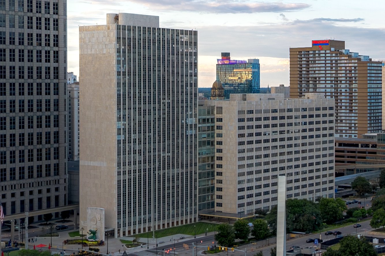 A tall modern civic building with a glass facade and plaza stands under a blue sky in downtown Detroit.