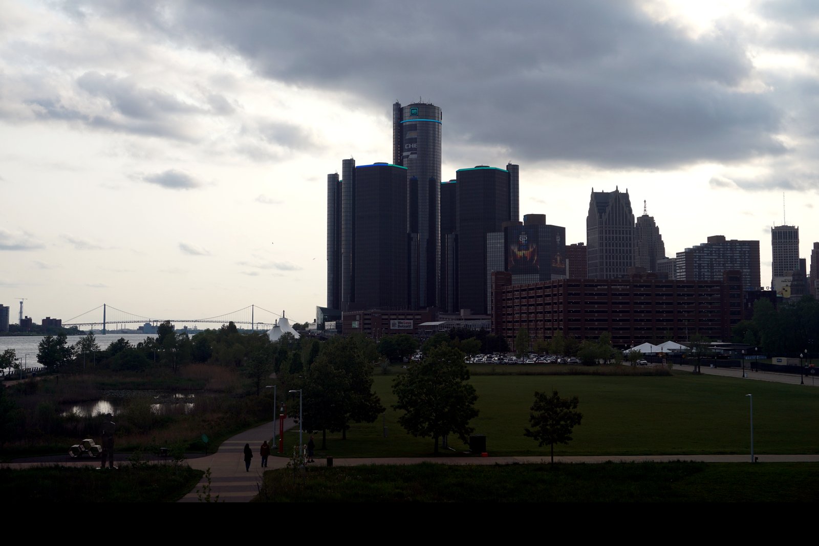The dark glass towers of the Renaissance Center rise above a wide green lawn and a riverside path under a cloudy sky, with the Ambassador Bridge visible far off to the left.