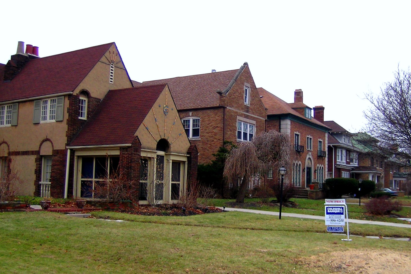 A row of large brick and stucco two-story houses with steep roofs and front lawns along a residential street.
