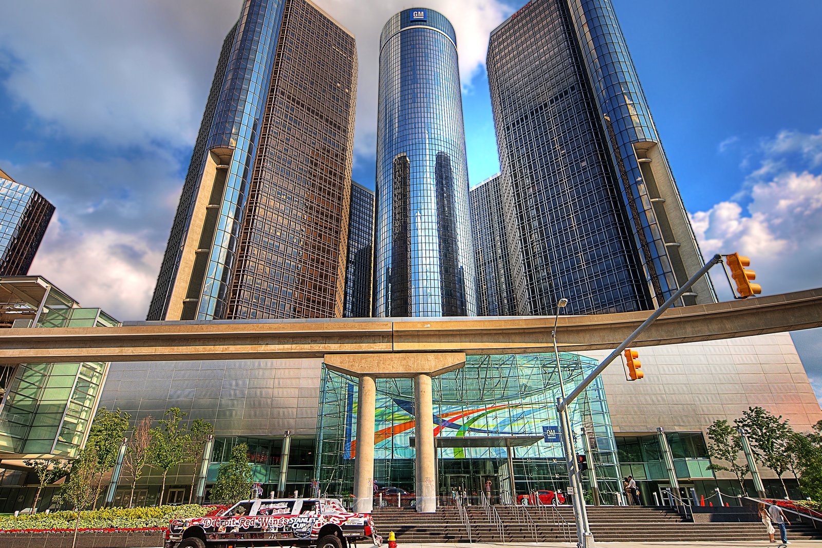 The glass towers of the GM Renaissance Center in Detroit seen from street level, with a skywalk and a red car in front.