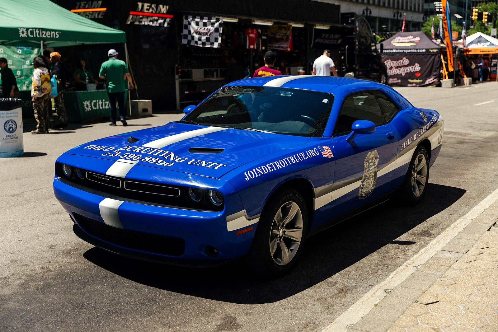 A blue and white Detroit Police Department field recruiting car, a Dodge Challenger, parked on a downtown Detroit street beside event tents.