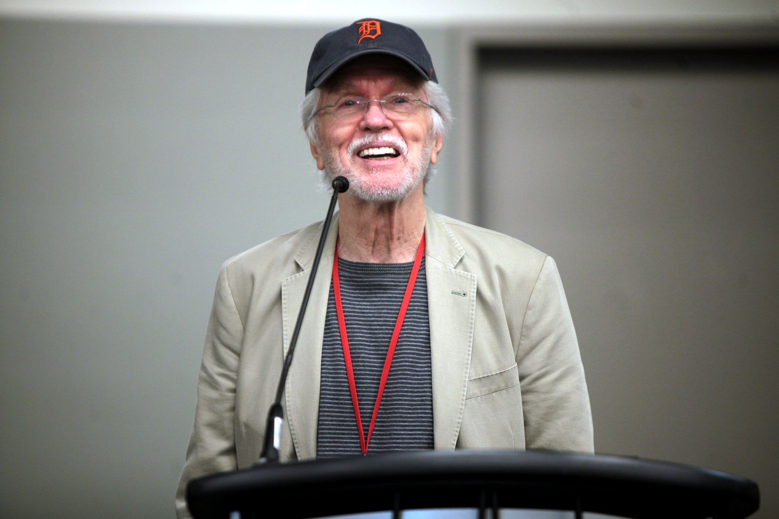 Tom Skerritt, smiling and wearing a Detroit Tigers cap, glasses and a tan jacket, speaks at a lectern with a microphone.