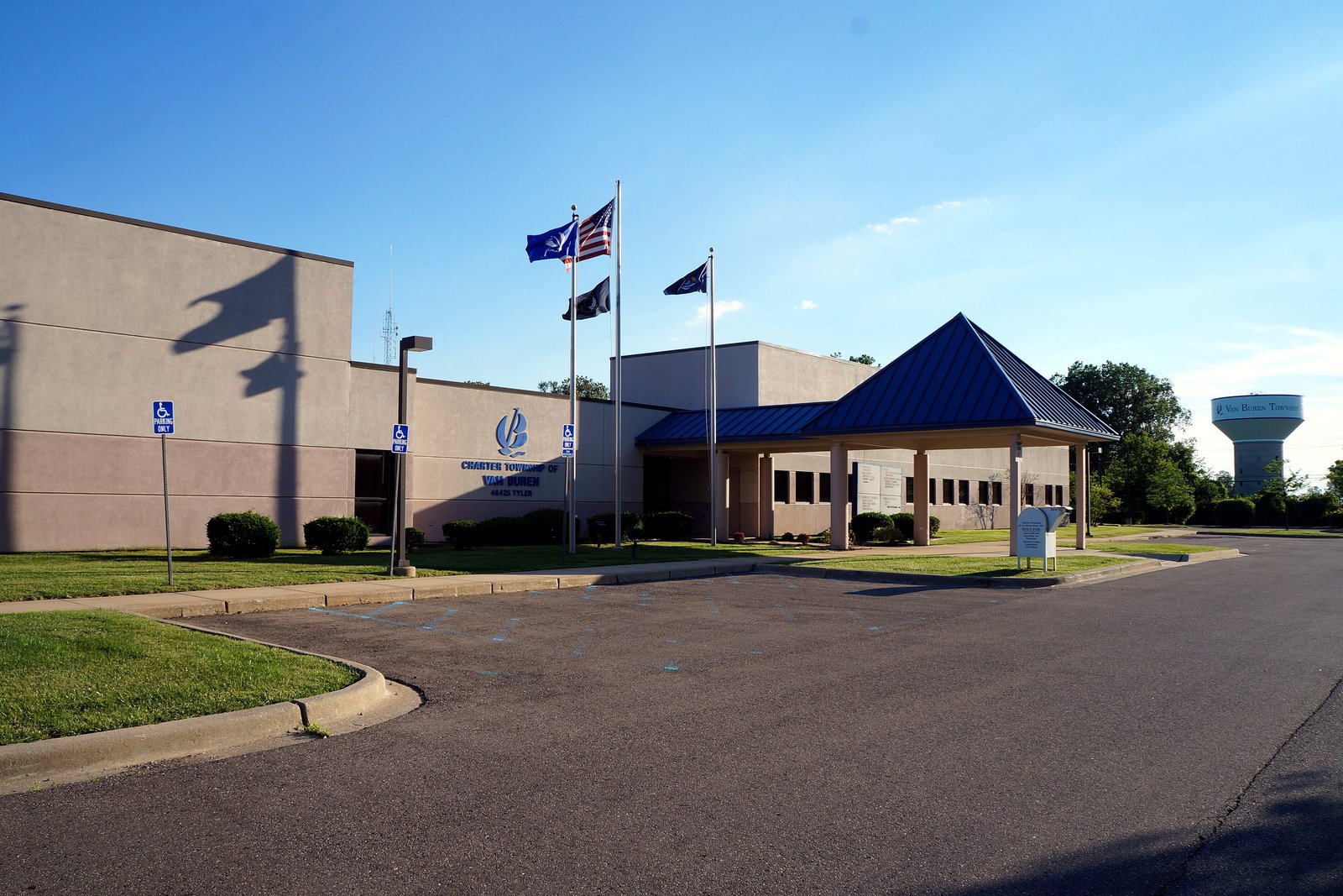 A low beige township hall with a blue peaked entrance canopy and flagpoles, with a water tower behind it under a blue sky.
