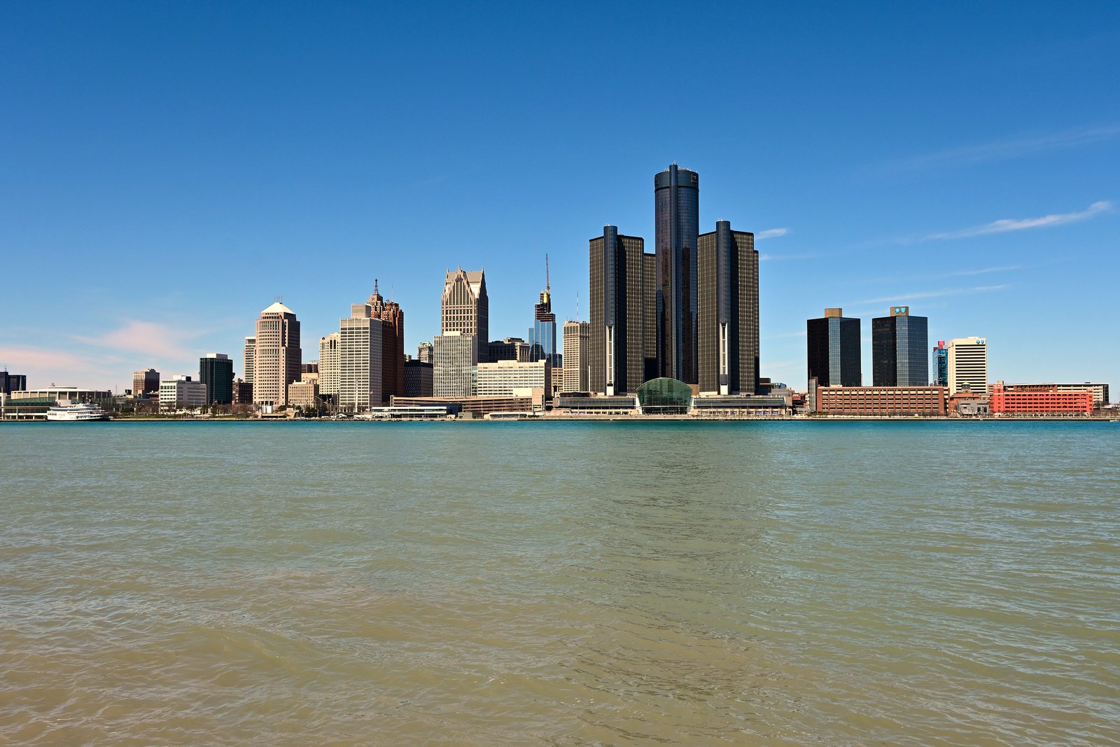 The Detroit skyline, led by the Renaissance Center towers, rises across the blue Detroit River under a clear sky, seen from the Canadian shore.