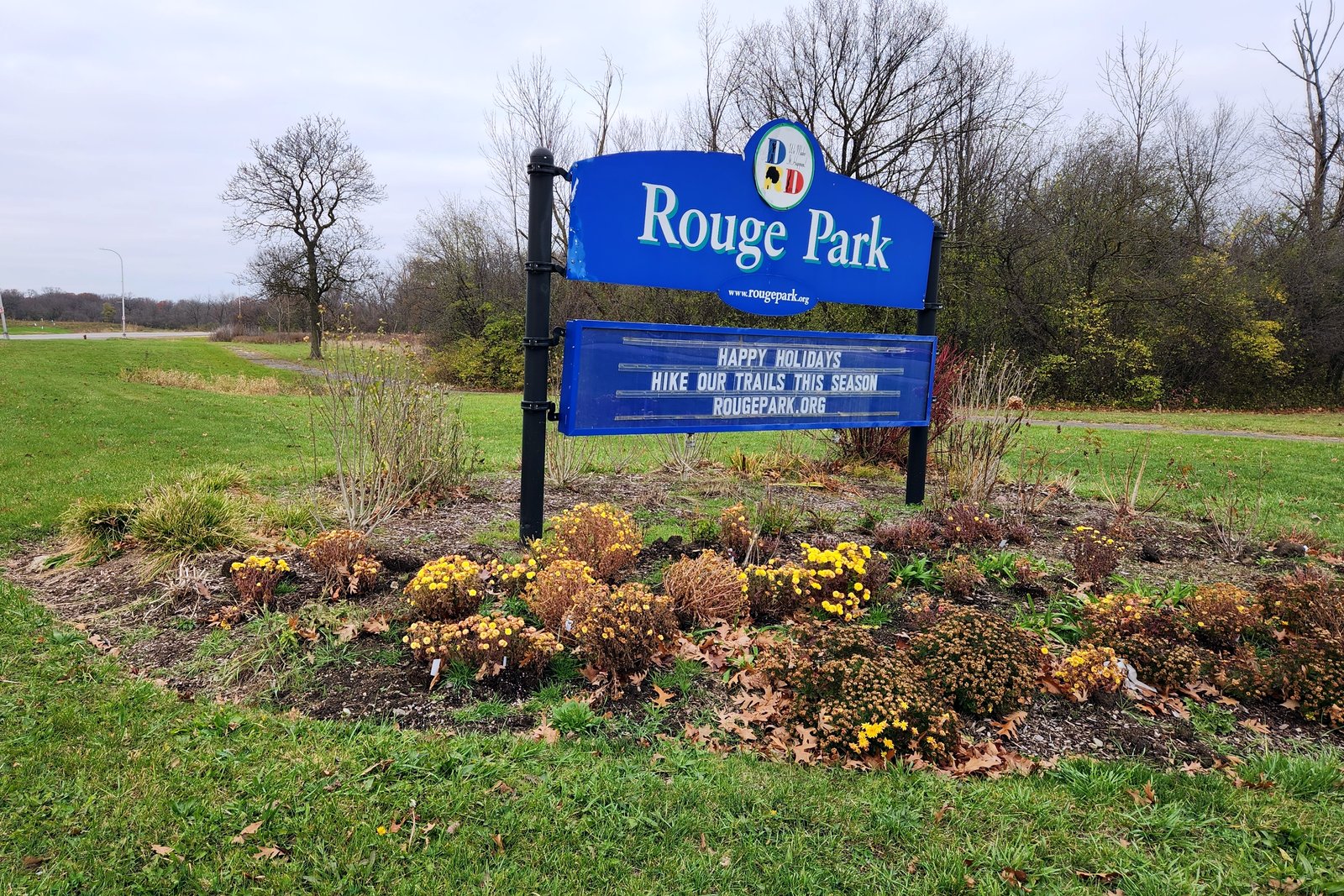 A blue Rouge Park sign stands in a bed of fading fall flowers beside a wide lawn, with bare trees under a gray sky.