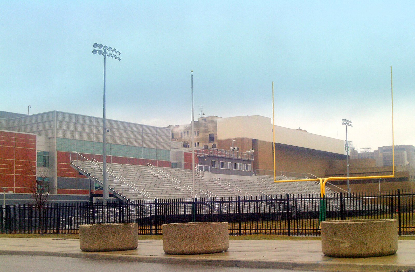 An empty football stadium behind a black iron fence, with tall gray bleachers, light towers and a yellow goalpost, in front of a large modern brick and metal school building under a hazy sky.