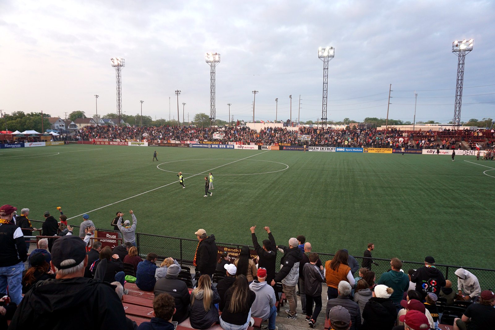 Fans stand along a railing at a soccer stadium as players line up on a green field under tall light towers.