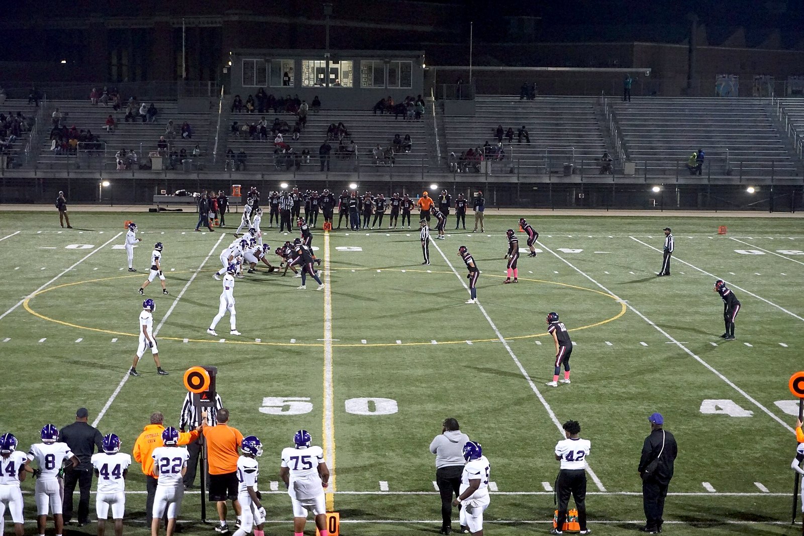 High school football teams line up for a play near midfield on an artificial-turf field at dusk as players and coaches watch from the sideline.