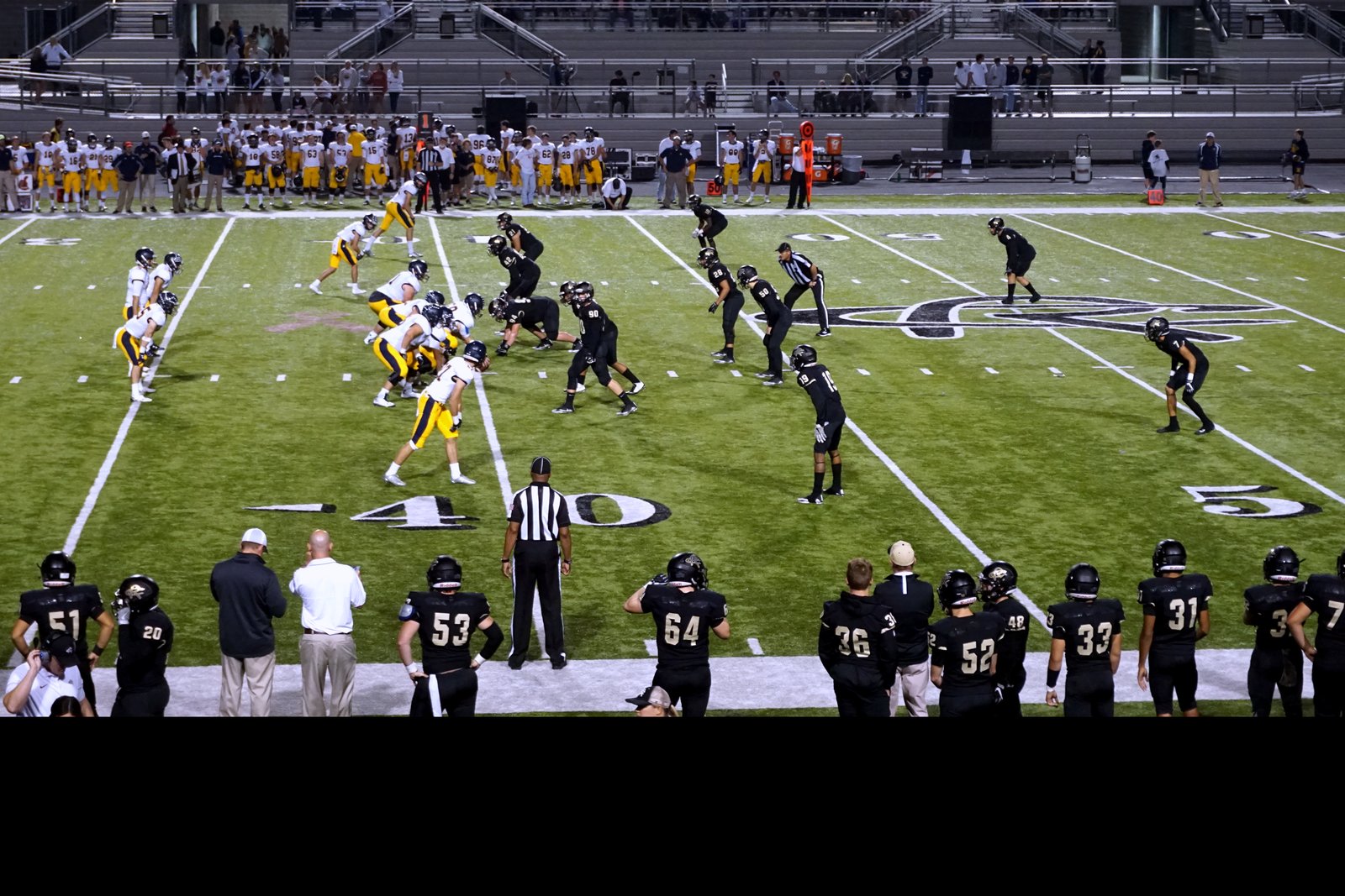 High school football players lined up at the line of scrimmage under stadium lights, with a crowd in the stands behind them.