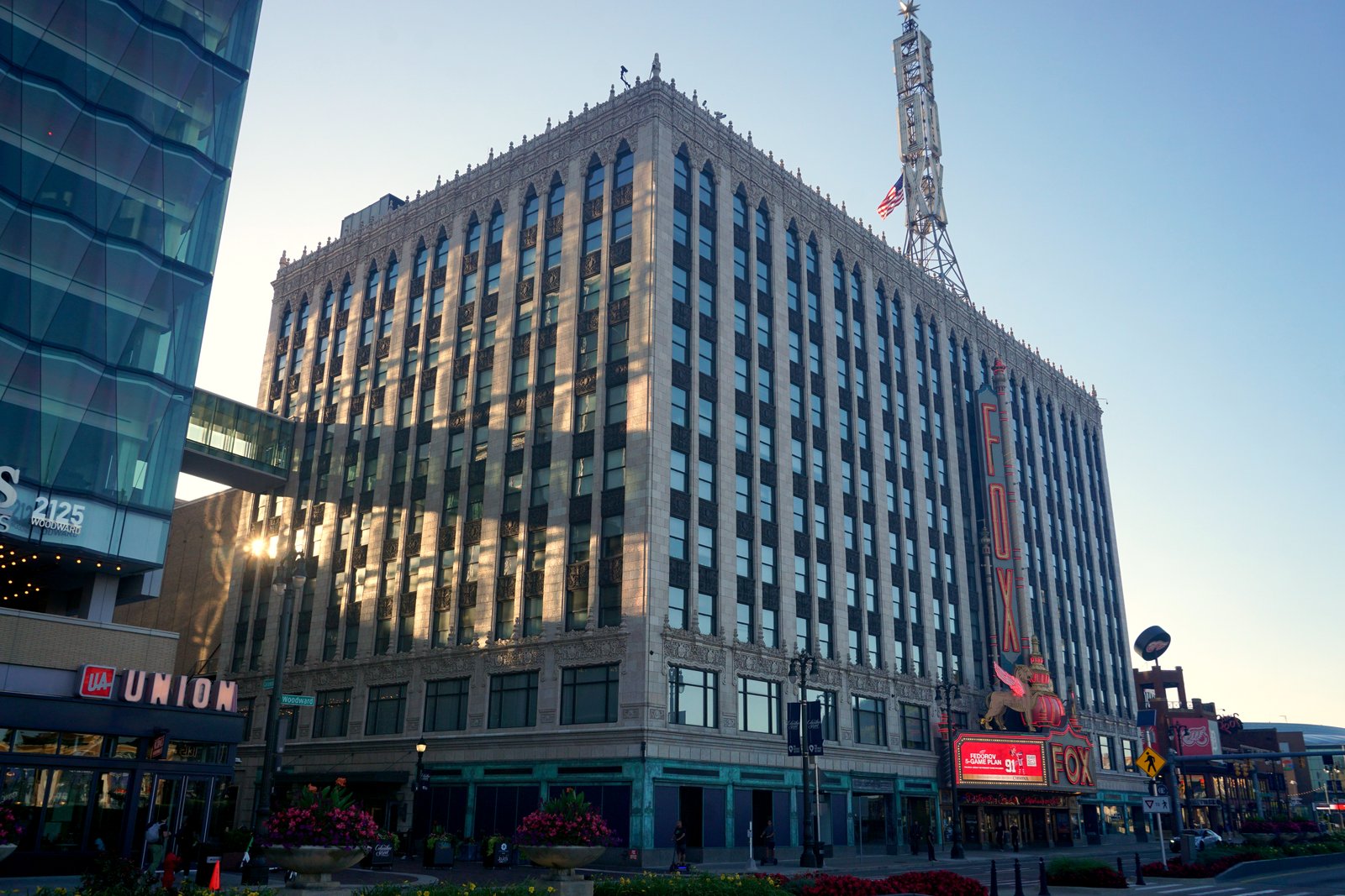 The tall, cream-colored Fox Theatre building on Woodward Avenue in Detroit, with its vertical red FOX sign and lighted marquee at the corner, under a clear evening sky.